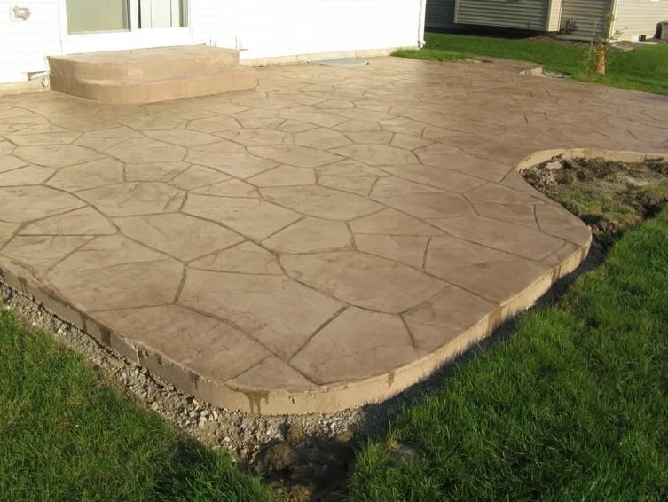 Stamped concrete patio in backyard with stone pattern design, bordered by green grass and gravel edge
