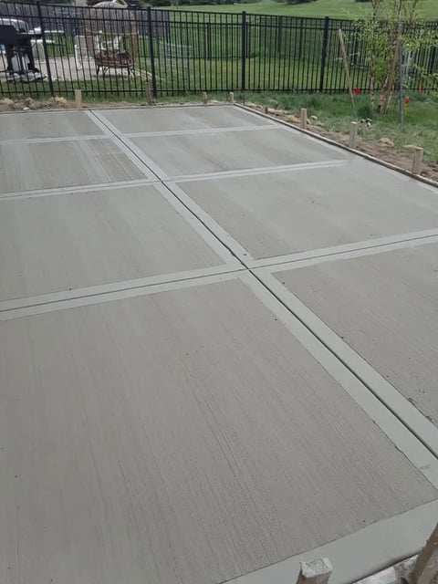 Concrete pool deck divided by white expansion joints with black metal fence and green grass in background