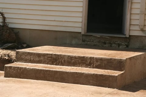 Two concrete steps leading up to a building entrance with white siding and a dark doorway in the background