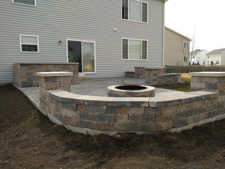 Backyard patio with curved stone retaining wall and central fire pit, adjacent to two-story house with gray siding