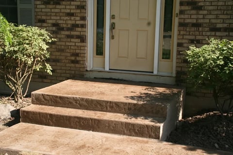 Concrete front entryway with two steps leading to a tan door on a brick house, flanked by green shrubs