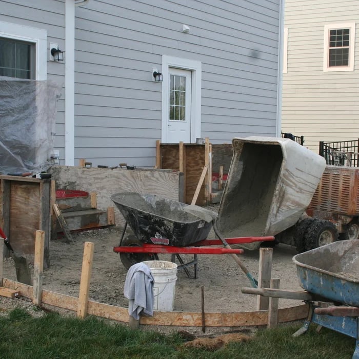 Construction area behind house with cement mixer, wheelbarrow, and building materials near wooden fence
